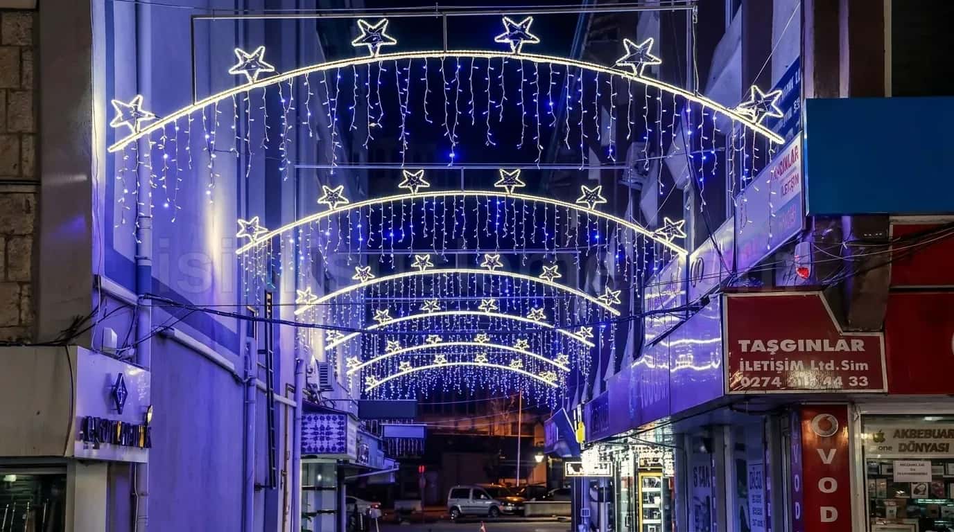 Gece cekilmis dar ticari sokakta yildizli mavi beyaz LED ark kemerleri ve bina arasindan gecen yayalar