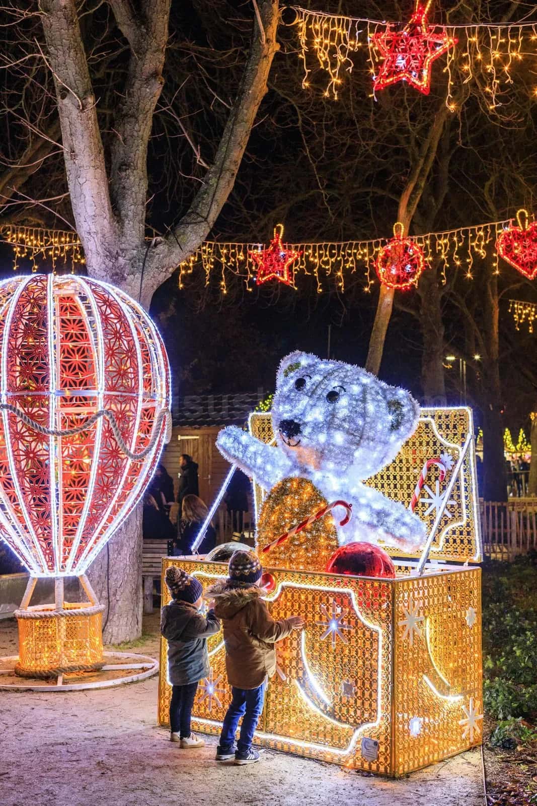 İki çocuğun dokunduğu LED polar ayı figürü ve kırmızı çizgili balon, arka planda kırmızı yıldız LED motifler