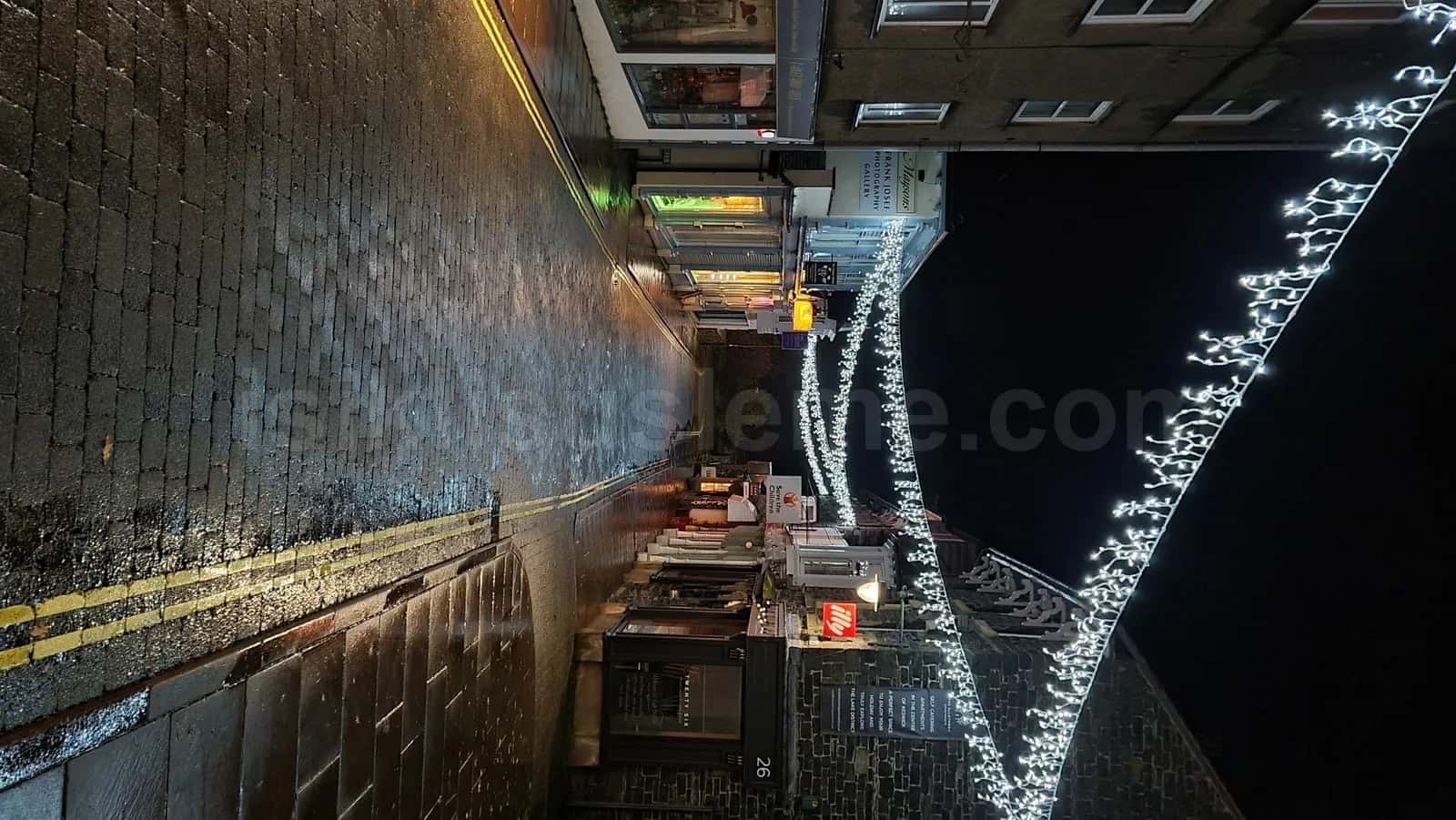 Islak tas kaldirim sokakta binalarin arasindan gerilen soguk beyaz LED zincir zig zag dizilimi gece gorunumu