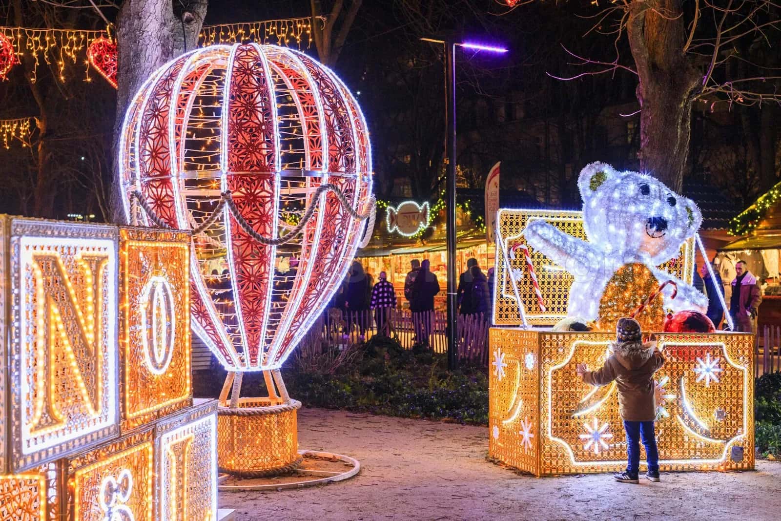 Yılbaşı parkında kırmızı beyaz LED balonun yanında kutu içinden çıkan beyaz LED polar ayı figürü önünde duran çocuk