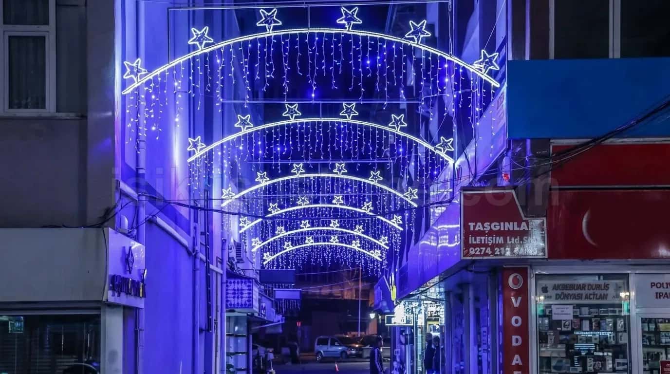 Dar ticari sokakta binalara bagli mavi beyaz yildizli LED ark kemerleri ve sarkacak LED perde bandi